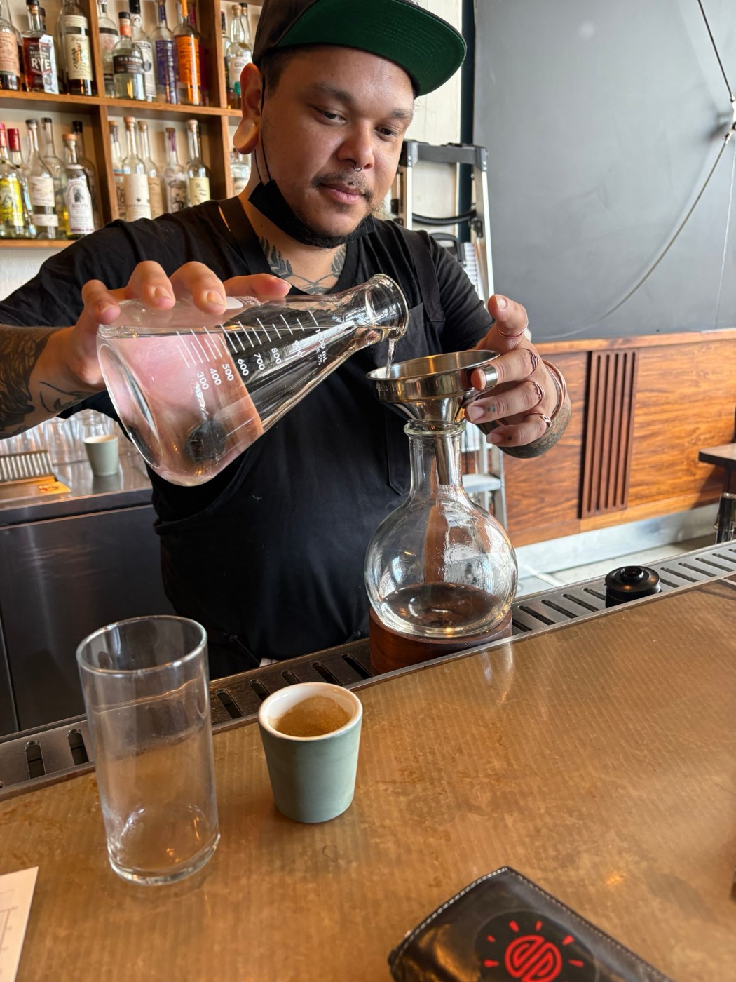 A bartender pours clear gin from a measuring flask through a funnel into a round bottle